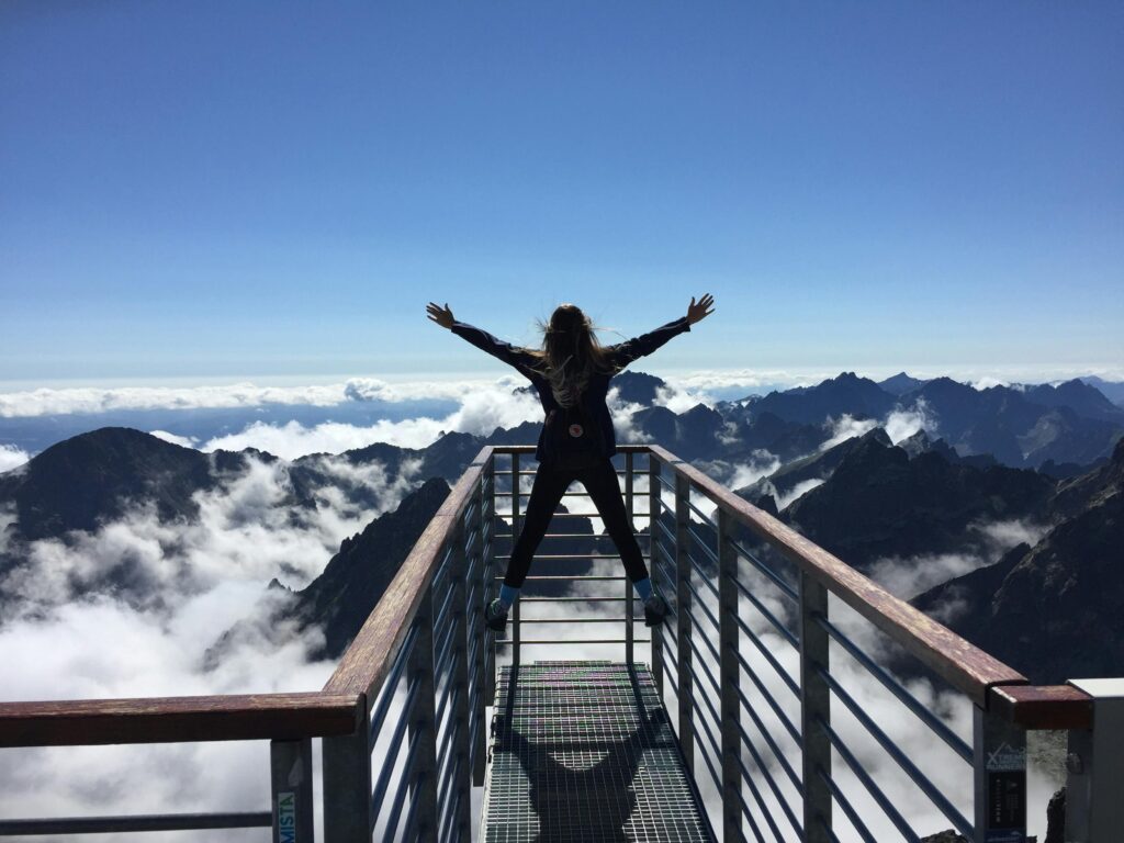 person standing on guard railing with arms open wide facing mountains